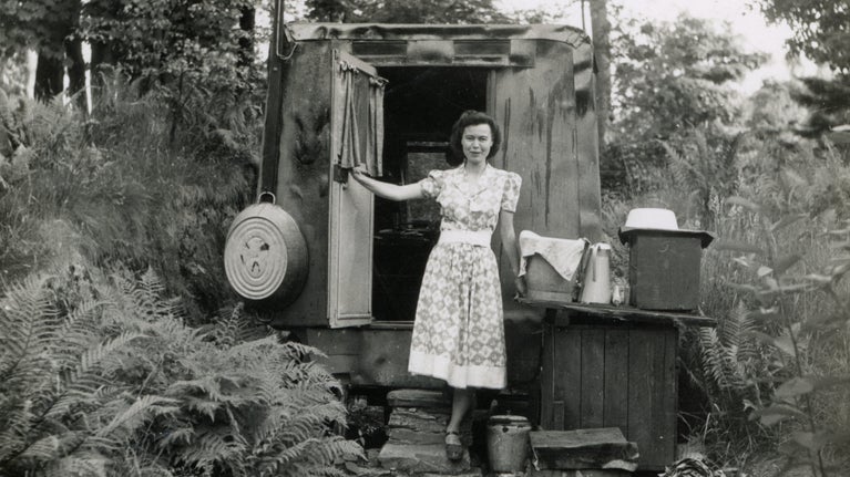 A vintage 1940s black and white photo of woman standing in front of an old caravan wearing a patterned day dress.